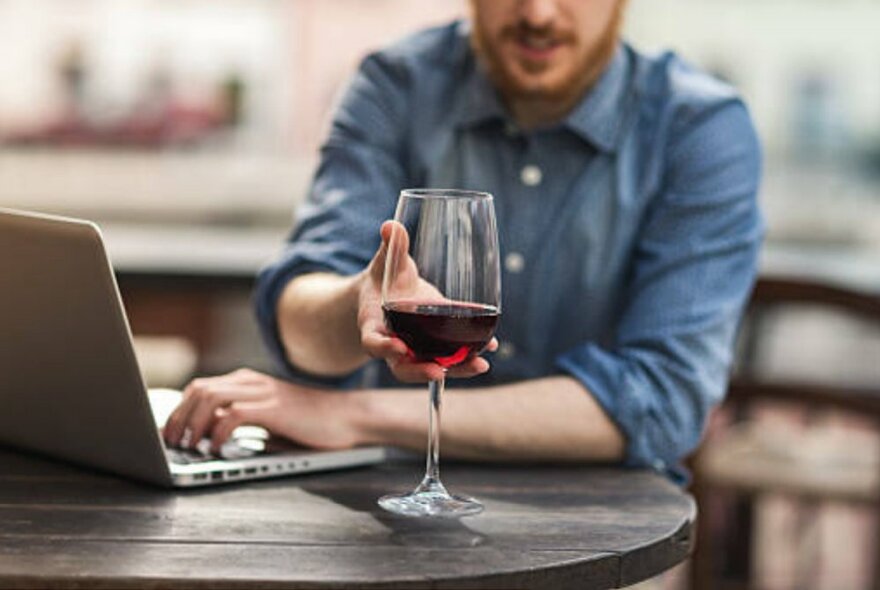 Person seated at a round table working on a laptop and reaching for a glass of red wine.