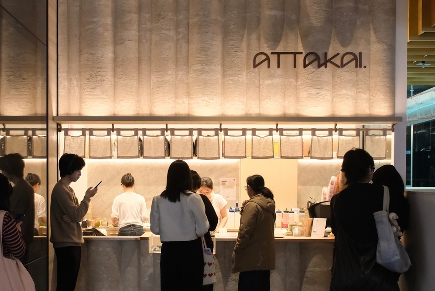 Queue at backlit matcha cafe counter, staff in white T-shirts preparing drinks.