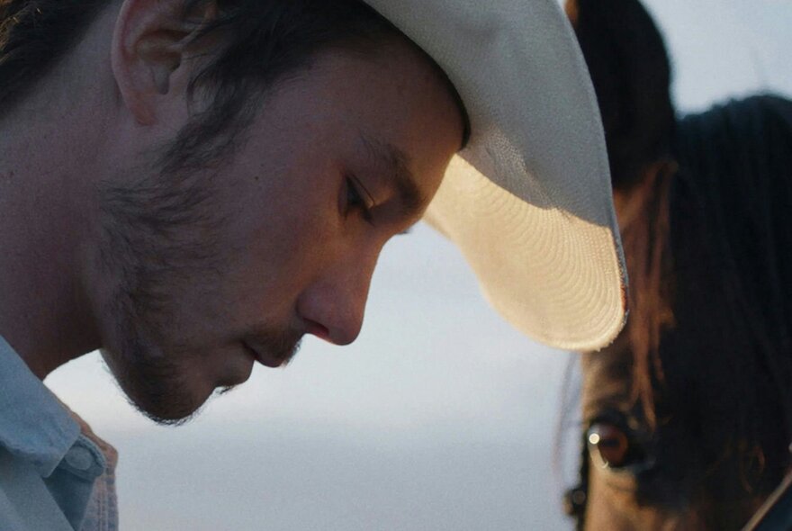 A still from the film The Rider; a profile of a young man wearing a cowboy hat, looking down, with part of a horse's head visible on the right.