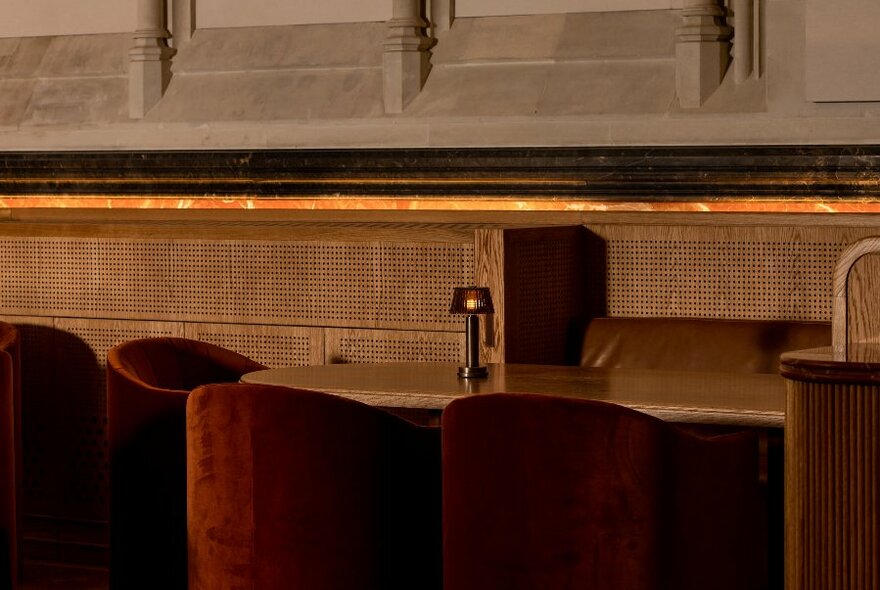 A dining table surrounded by dark rust coloured velvet club chairs, positioned near a wall with details cut into the stone wall plinths.