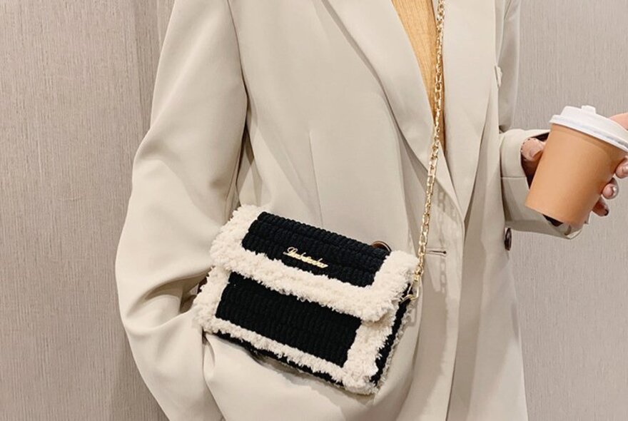 A person modelling a black and white knitted shoulder bag with plush trim and a chain strap, made in a cross-stitch workshop.