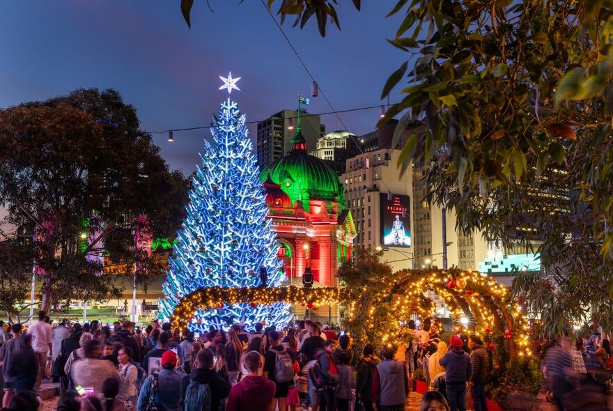 A large crowd of people around the giant illuminated Christmas tree at Fed Square, other lit up city buildings in the background; night time.