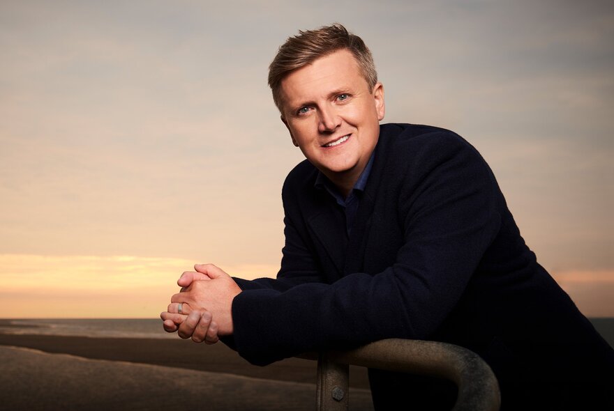 Aled Jones, wearing a dark jumper and leaning forward over a railing, his hands clasped, with a twilight sky behind him.