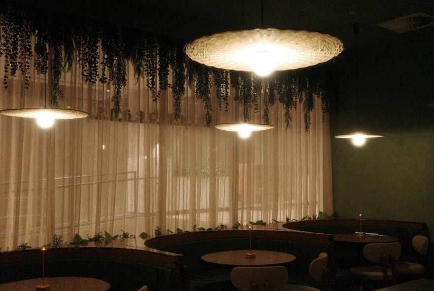 Interior view of a dimly lit bar or restaurant with booth seating, round tables, and hanging pendant lights over white curtains and foliage decorations.