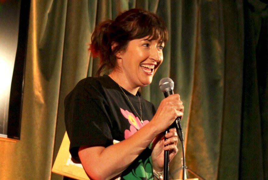 Comedian Celia Pacquola talking into a microphone she holds in her hand,  performing live on stage.