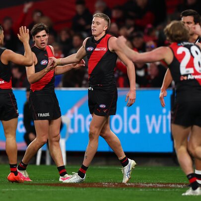 A huddle of Essendon AFL players celebrating on the field after having scored, with blurred fans in the stadium in the background.