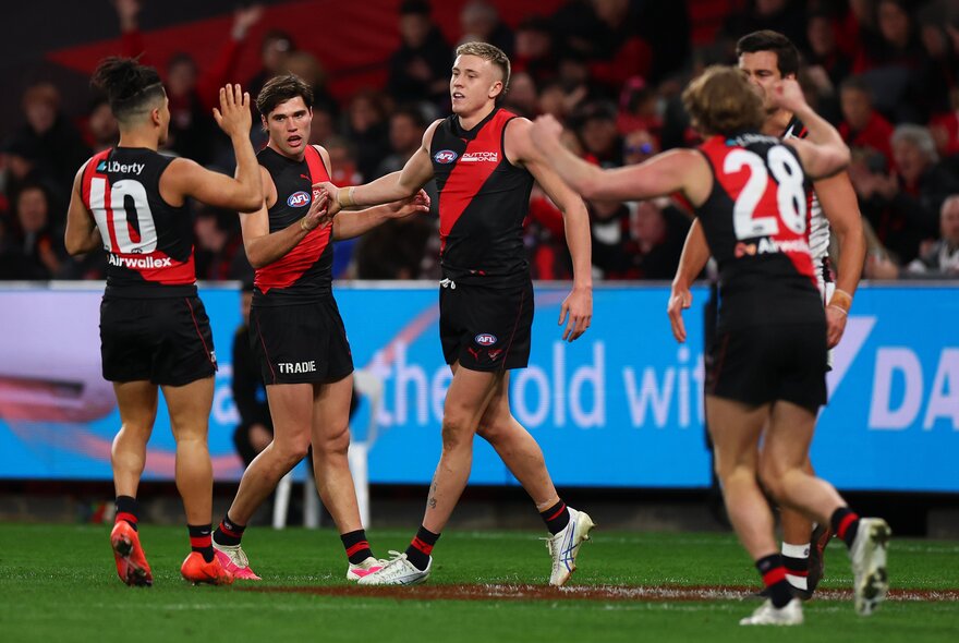 A huddle of Essendon AFL players celebrating on the field after having scored, with blurred fans in the stadium in the background.