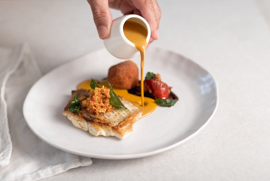 A hand pouring a small pot of sauce over a fish fillet at a fine dining restaurant. 