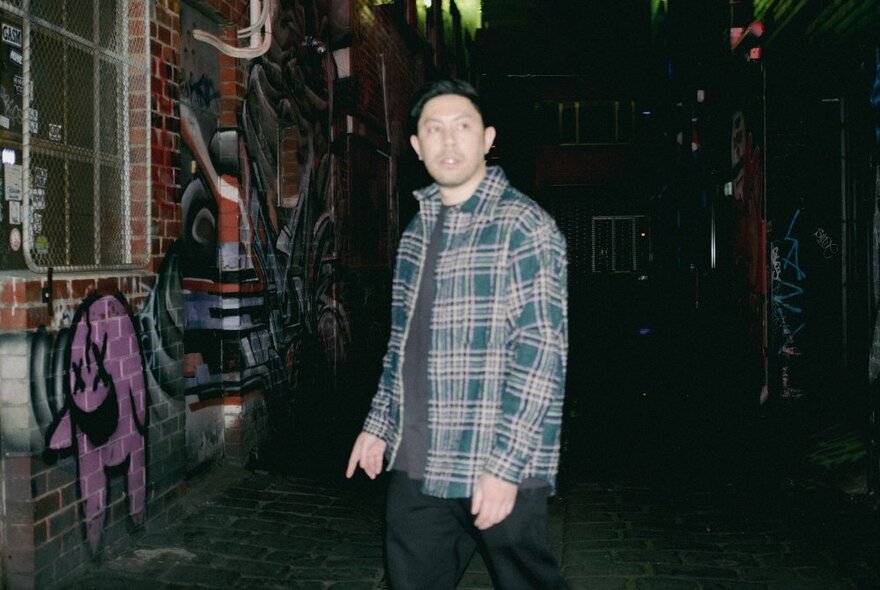 Comedian Chris Nguyen, a young man standing outside a building at night time, wearing a flannelette shirt and dark trousers.