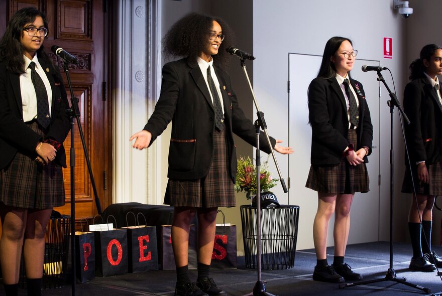 Four girls in school uniform stand on stage behind microphones, smiling. 