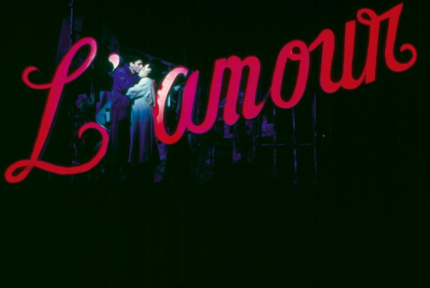A couple on stage kissing in front of a red cursive sign saying L'amour.