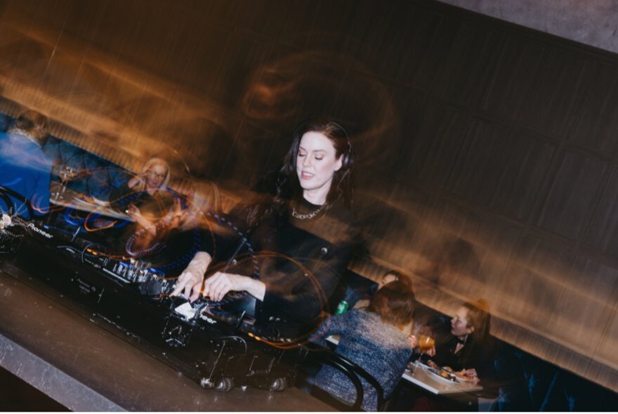 A DJ mixing the controls at a DJ booth, behind her are patrons seated at tables dining.