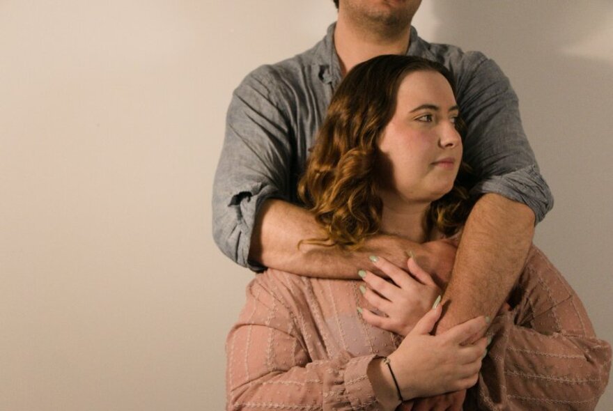 A woman in a pink blouse looking off to her left, being hugged from behind by a man in a grey shirt in front of a beige background. 