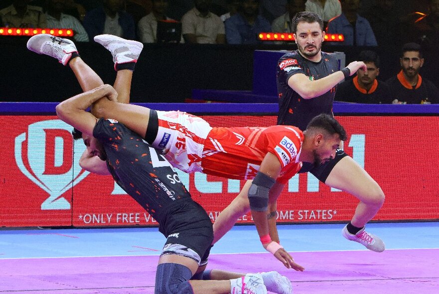 Three players wearing protective elbow and knee pads tackling each other in the sport of kabaddi, in an indoor sporting arena.