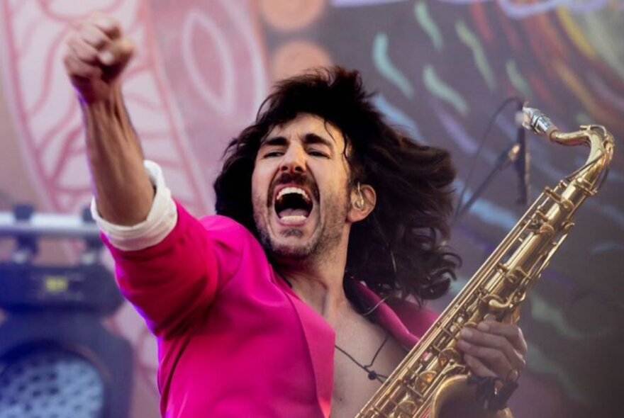 A saxophonist on stage wearing a pink blazer and pumping his fist while holding his saxophone.