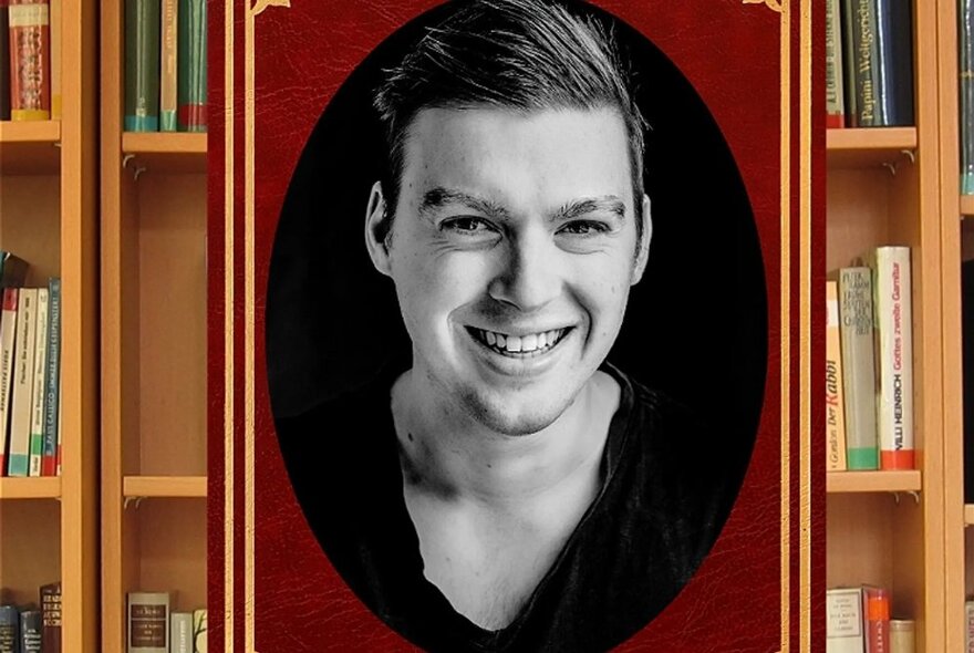 Head and shoulders portrait of a smiling man, set in an oval frames, in front of library bookshelves filled with books.