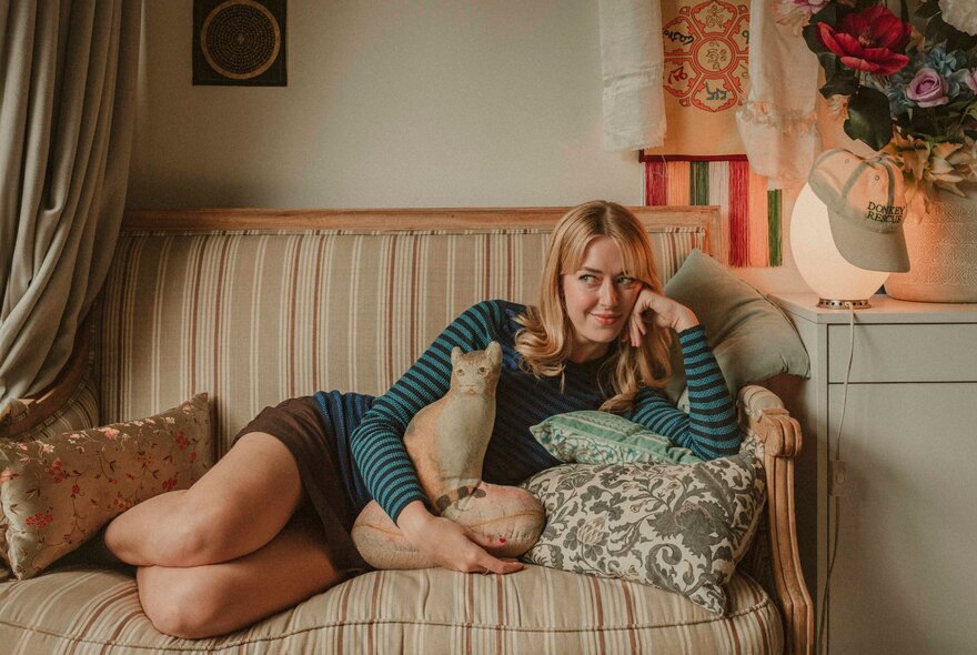 Artist and creator Celeste Mountjoy, lying on a small couch, her head propped up by her arm, leaning on soft cushions, a lamp and artwork next to her.