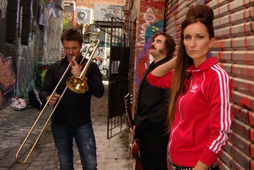 Band in a back alley with graffiti, woman wearing sports top with long wig in the fashion of Amy Winehouse, one musician with a trombone.