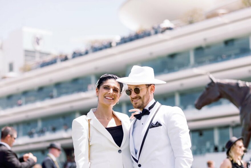 Two well-dressed people posing with a large building behind them and a bronze statue of Makybe Diva. 