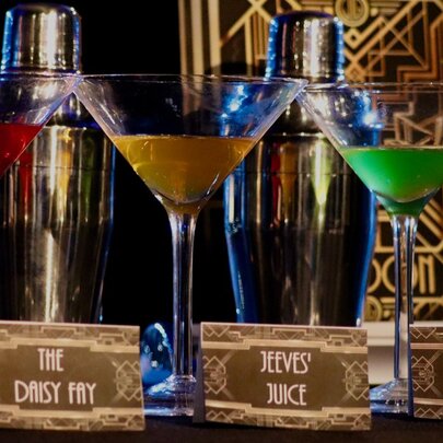Three martini glasses each with a different coloured liquid in it, and art deco style name cards in front of them.