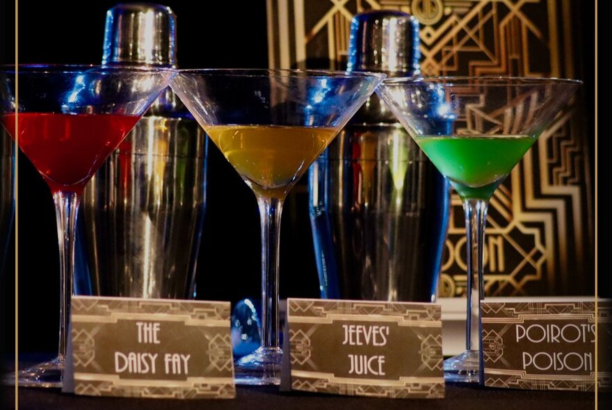 Three martini glasses each with a different coloured liquid in it, and art deco style name cards in front of them.