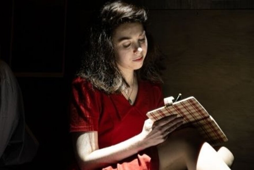 Actor portraying Anne Frank, writing in her diary in a darkened room.