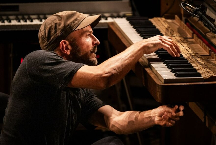 German composer and musician, Nils Frahm, crouching in front of a piano keyboard with one hand above, playing the keys, and one hand below, holding on to something.