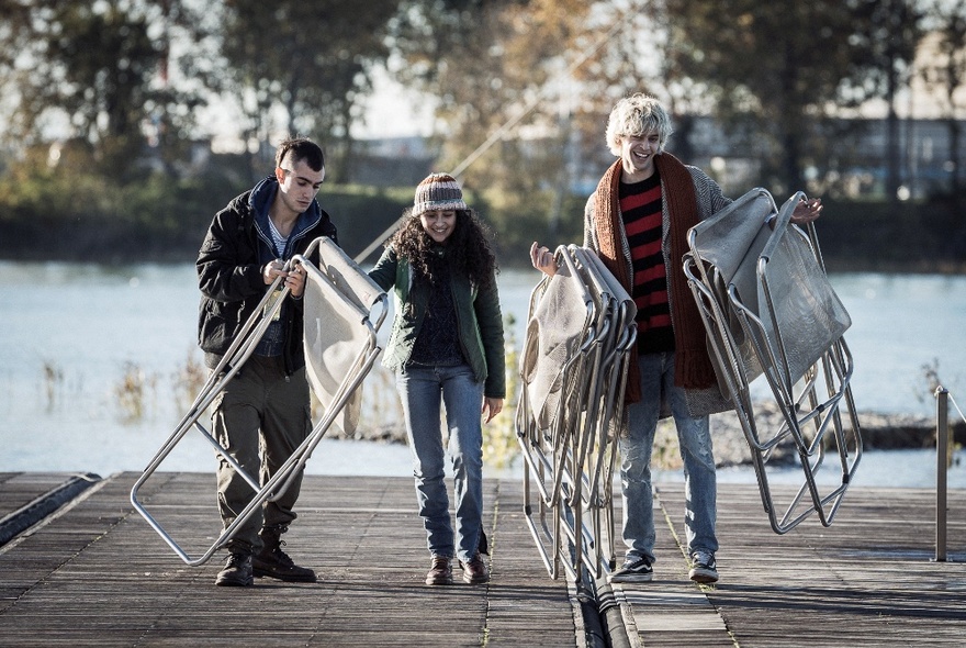 A still image from a film showing three people by a lake stacking deckchairs.
