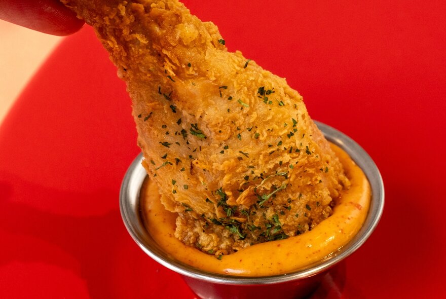 A Korean fried chicken drumstick being dipped into a small silver bowl of dipping sauce, against a red background.