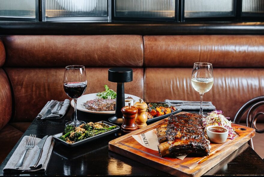 A booth in a restaurant, with a wooden tray of cooked steak, glasses of wine and other dishes of food arranged on the table.