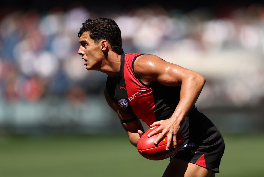 An Essendon AFL player holds the ball and crouches over slightly on the run. 
