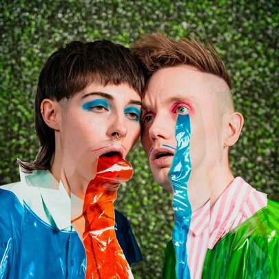 Two creative performers with their foreheads pressed together, both wearing plastic over their clothes and one with red plastic coming from his mouth and the other with blue plastic coming from his eye.
