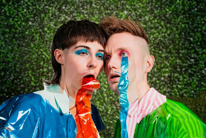 Two creative performers with their foreheads pressed together, both wearing plastic over their clothes and one with red plastic coming from his mouth and the other with blue plastic coming from his eye.