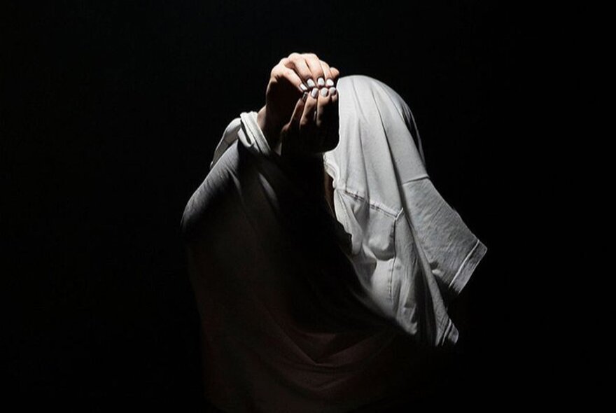 Physical theatre performer with his hands cupped together, a white t-shirt stretched over his hunched body, on a dark stage.