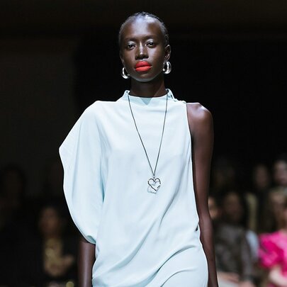A fashion model wearing a very pale blue, asymmetrical draped dress with a sculptural voluminous sleeve detail on a runway.