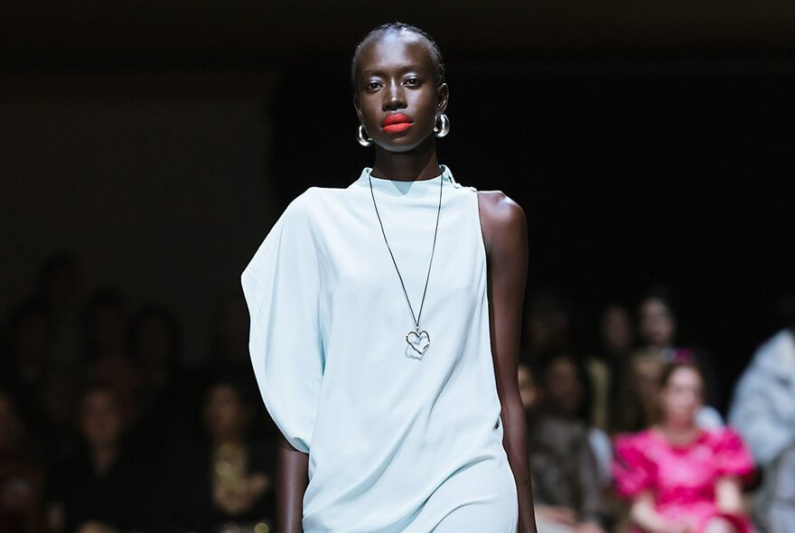 A fashion model wearing a very pale blue, asymmetrical draped dress with a sculptural voluminous sleeve detail on a runway.