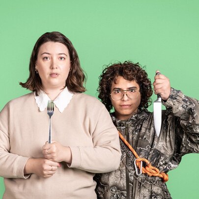 Two individuals posing in front of a solid green background, one holding a fork, the other holding a chef's knife. 