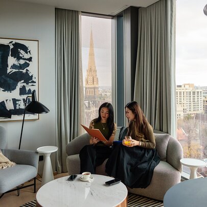 Two friends are reading a book in a modern hotel room. There is a view of Melbourne city behind them.