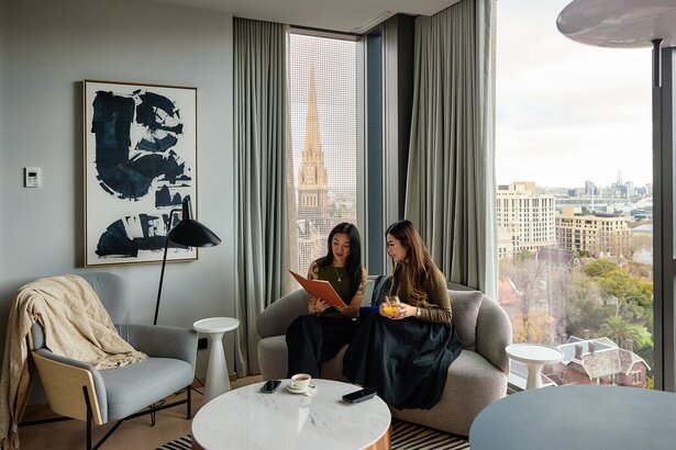 Two friends are reading a book in a modern hotel room. There is a view of Melbourne city behind them.