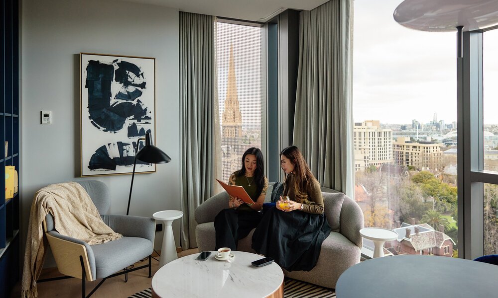Two friends are reading a book in a modern hotel room. There is a view of Melbourne city behind them.