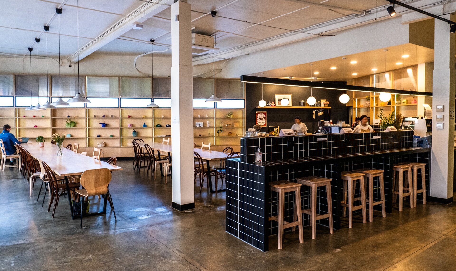 Inside a modern cafe. There are people making coffees at the counter and a shelf with ceramics at the back of the room.