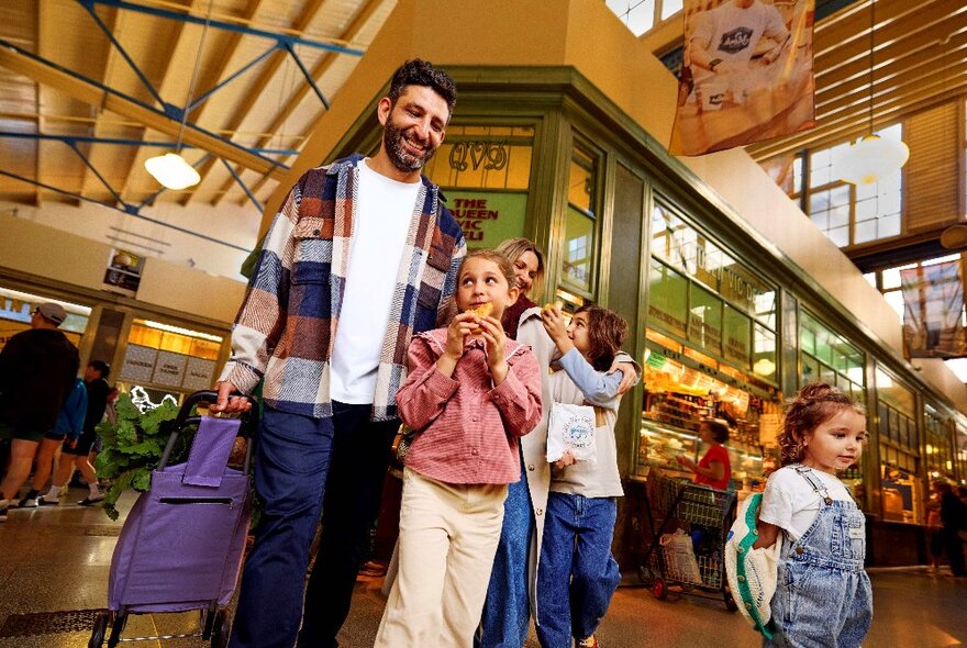Father and three children walking through Queen Victoria Market Deli hall with shopping and snacks.