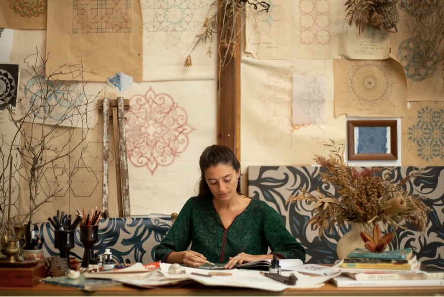 Woman seated at a table working with paper and crafts materials, mandala artworks on the wall behind her.