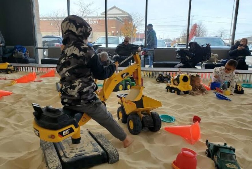 A child plays on a digger in a large sandpit filled with toys in front of a large window. 