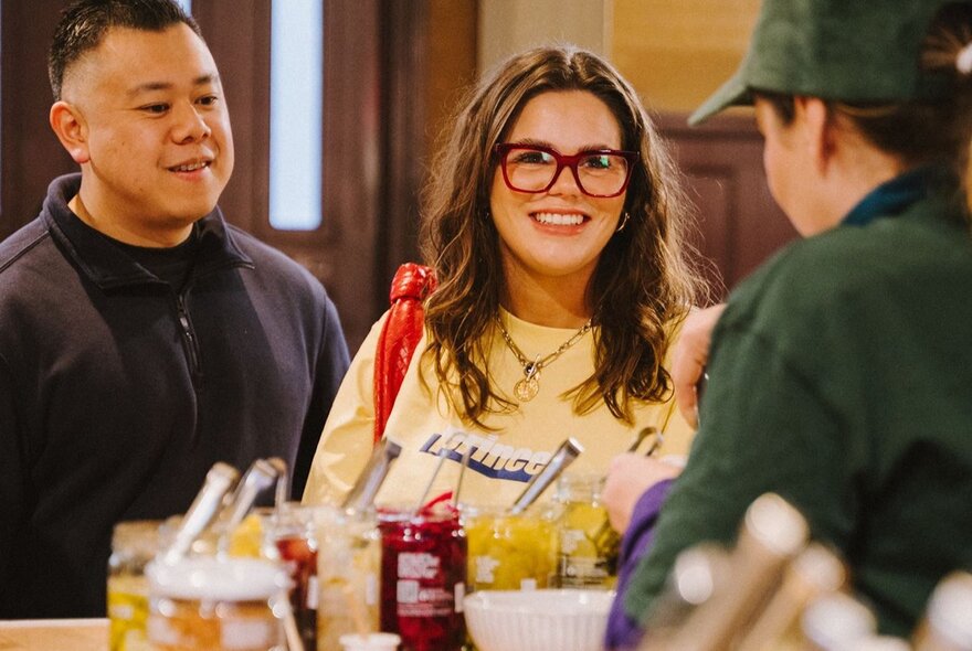 Two people interacting with a stall holder displaying condiments at an indoor market. 