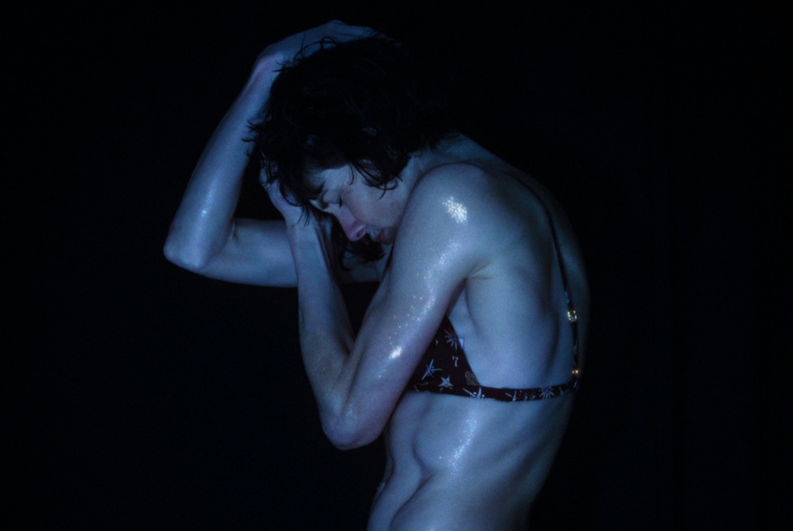 A dark image of a wet woman in a bikini top curling her body with her arms on clasping her head. 