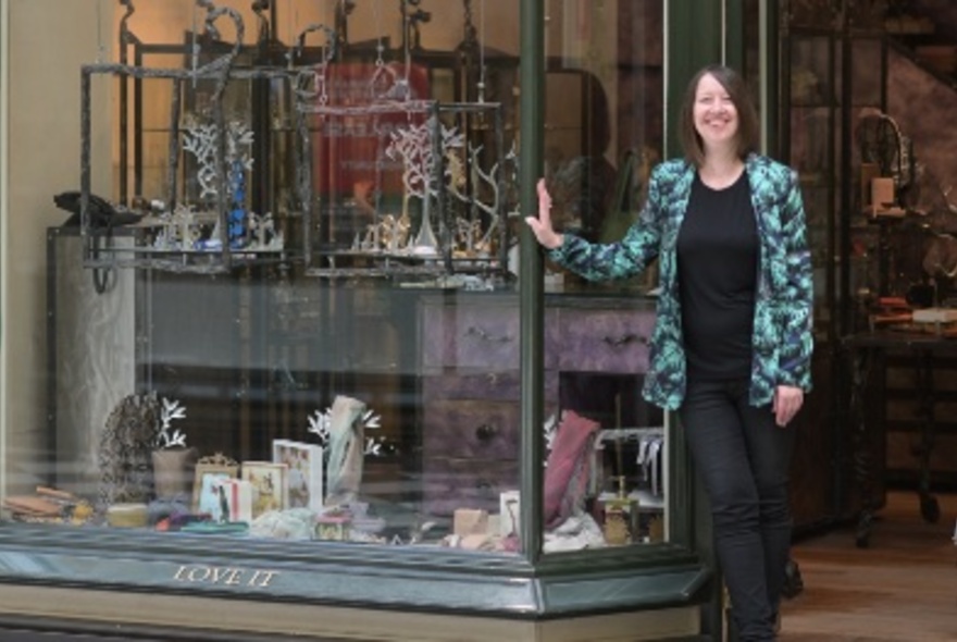 Smiling woman standing at the entrance to the shop Love It with her hand resting on the large glass window.