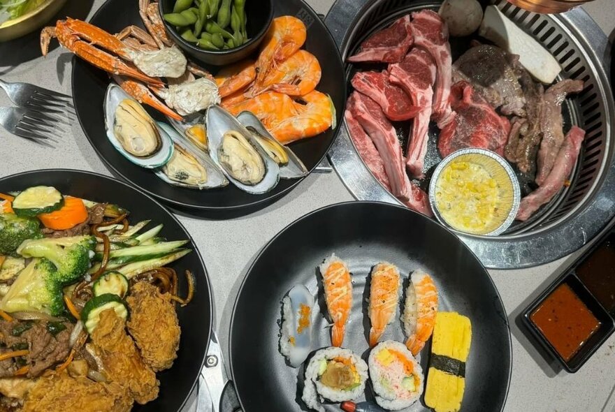 Four dishes of Korean food including ready to cook chops, oysters and seafood, sushi and vegetables.