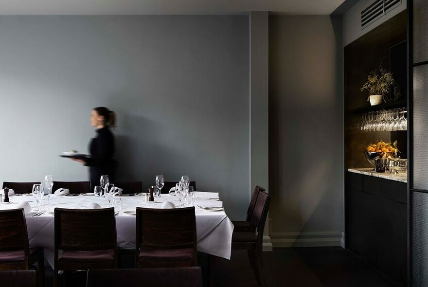 Waitress carrying a tray past a long dining table with white napery.