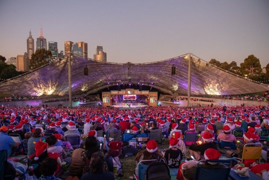 A crowd on people sitting on the grass at the Sidney Myer Music Bowl wearing Santa hats.